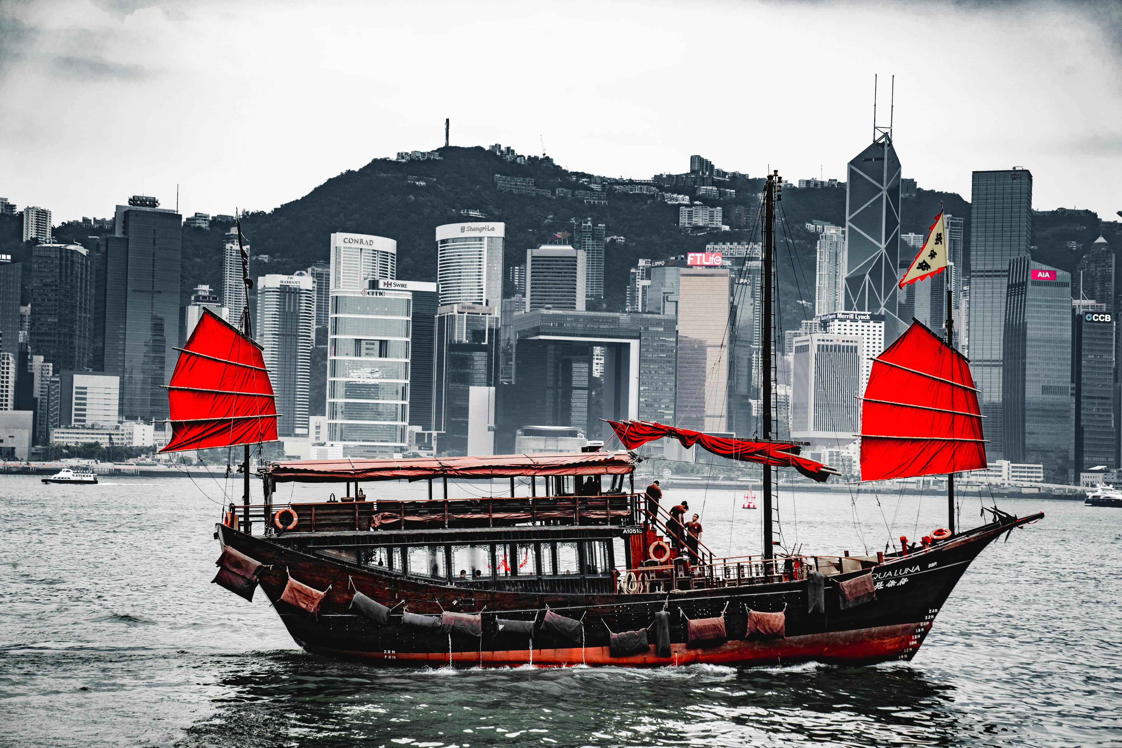 A traditional junk boat with red sails in Hong Kong harbor, against a skyline backdrop.