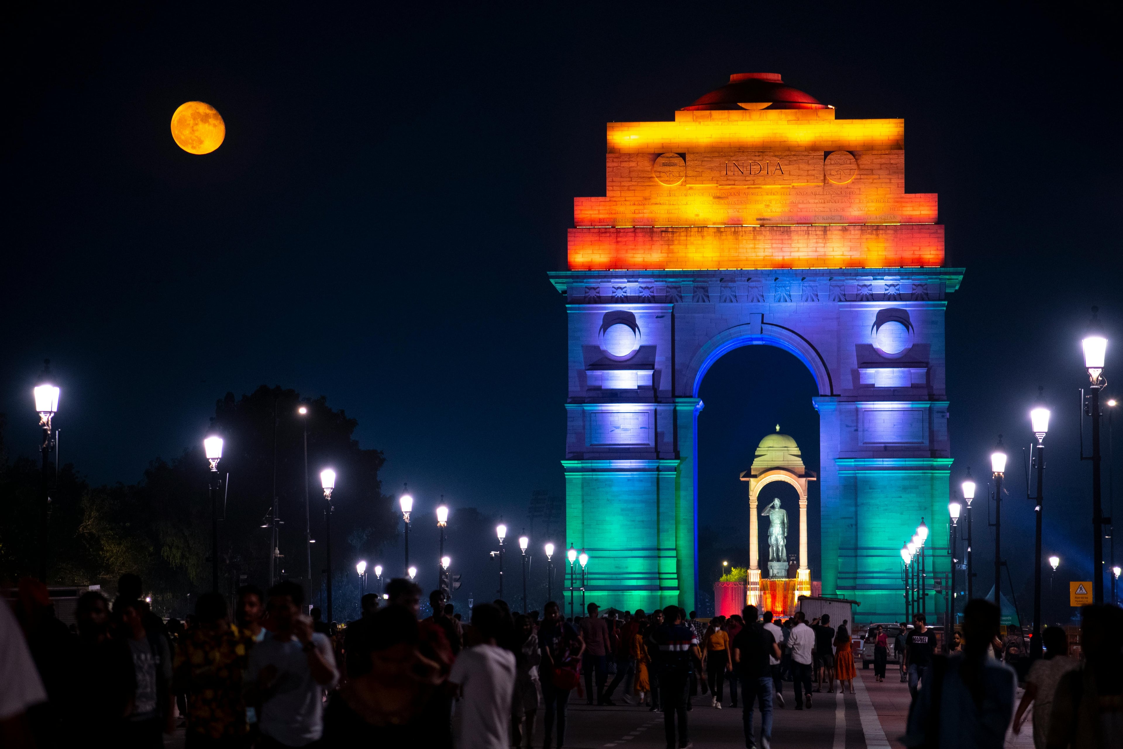 A lively night scene at India Gate in New Delhi, illuminated with colorful lights under a full moon.